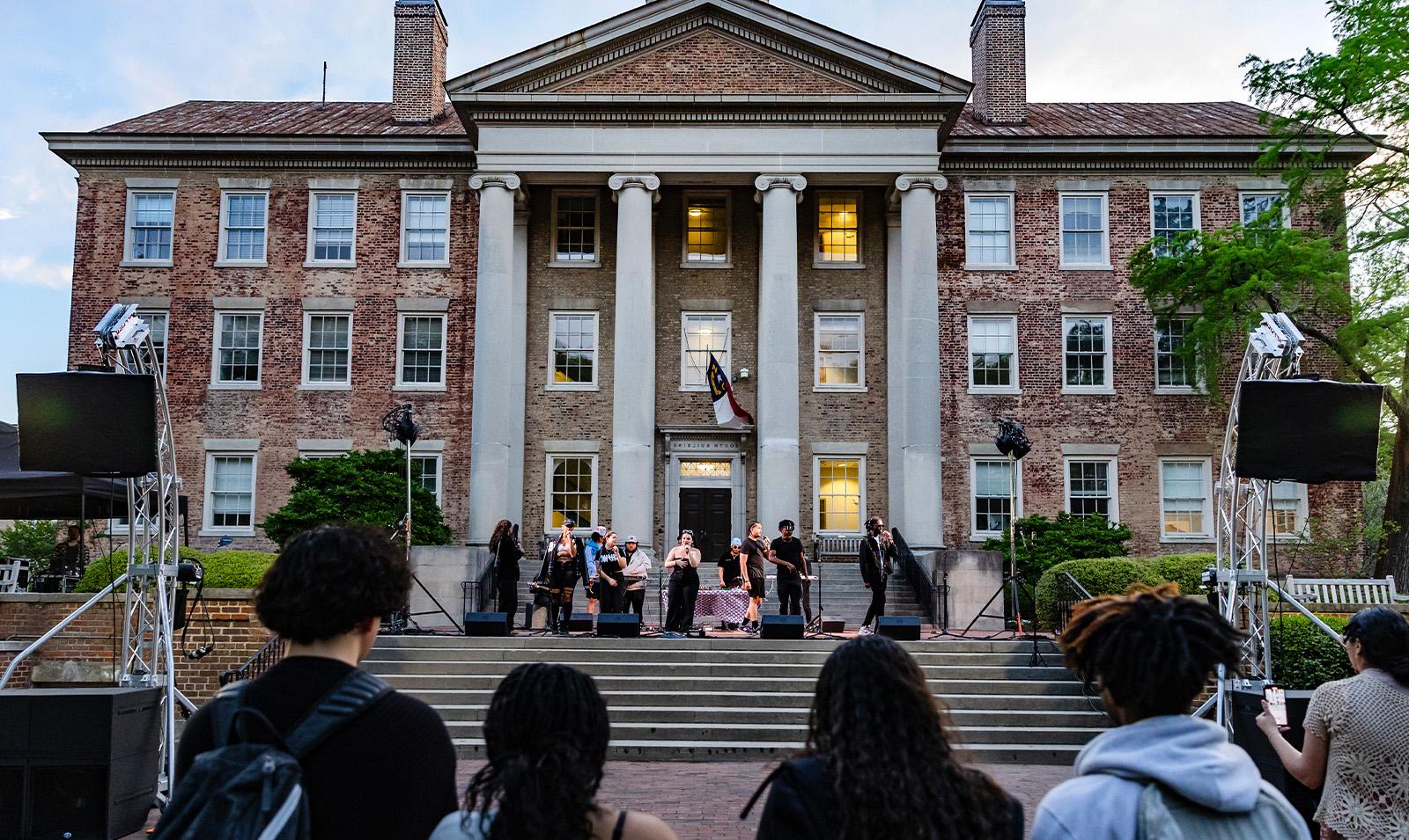 A band performing on the steps of South Building. In the foreground are students watching.